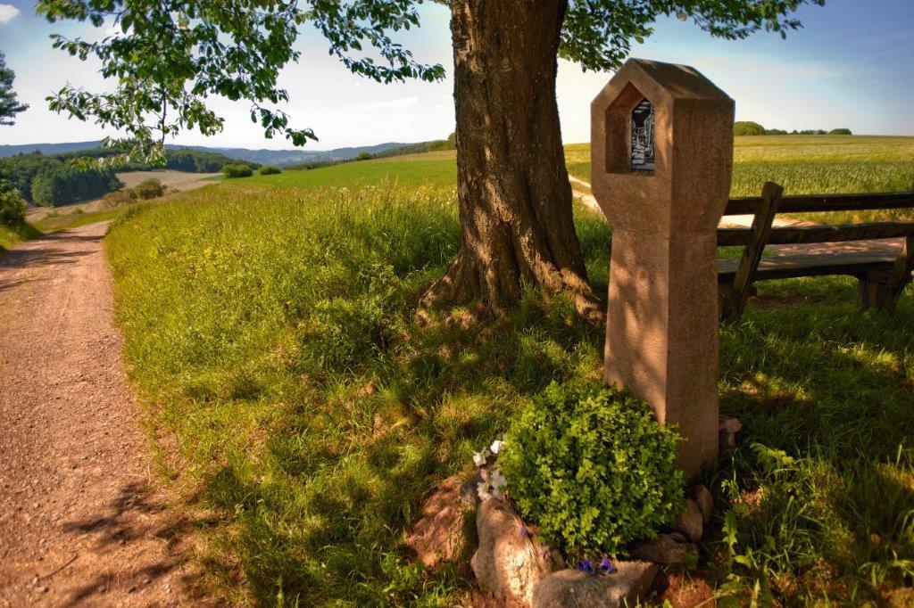 Der Bildstock aus Sandstein steht in der Nähe eine Baumes und einer Bank
