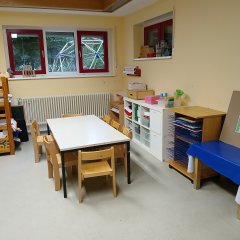 In dem Zimmer steht ein Tisch mit 6 Stühlen, diverses Bastelmaterial wie Farben und Papier ist zu sehen. Aus dem Fenster kann man das Klettergerüst im Garten sehen