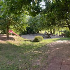 Blick in den Garten des Kindergartens Purzelbär mit Klettergerüst, Sandkasten, Schaukel und vielen Bäumen die Schatten spenden im Sommer